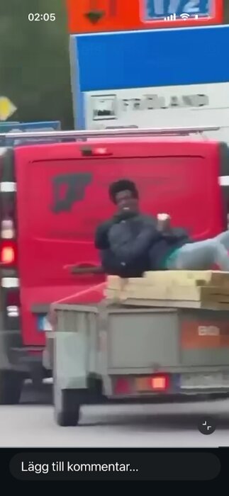 En person ligger på plankor som är fastsurrade på en släpvagn, dragen av en röd bil på en väg.