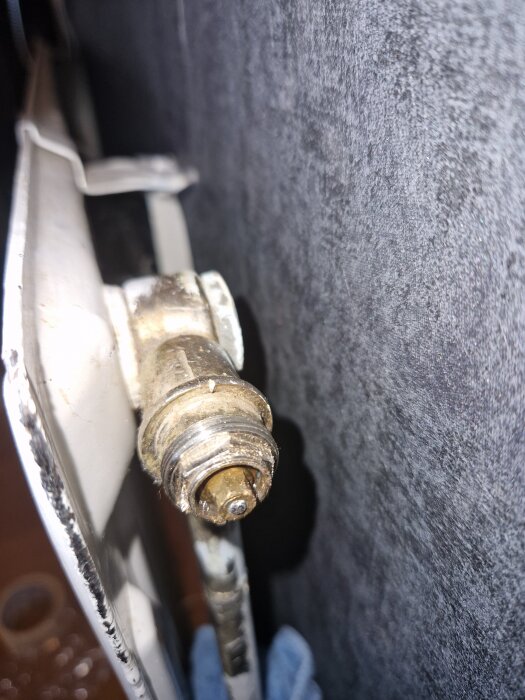 Närbild av en gammal radiatorventil monterad på ett element, med detaljer och viss rosthärjad yta mot en mörk bakgrund.