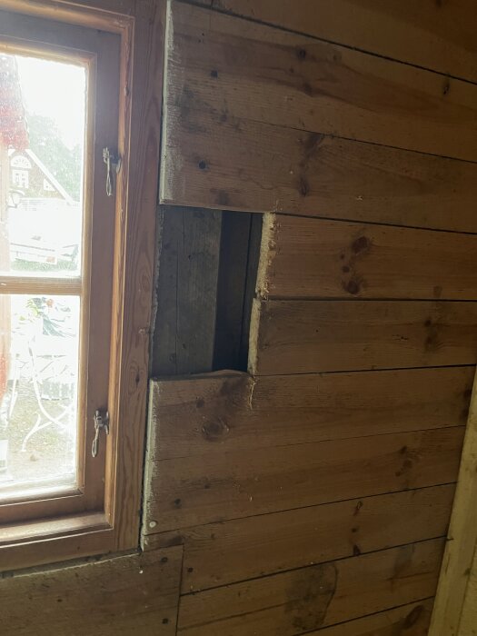 Träväggen har en öppning bredvid ett fönster med karm av trä, där några brädor saknas och isolering är synlig.