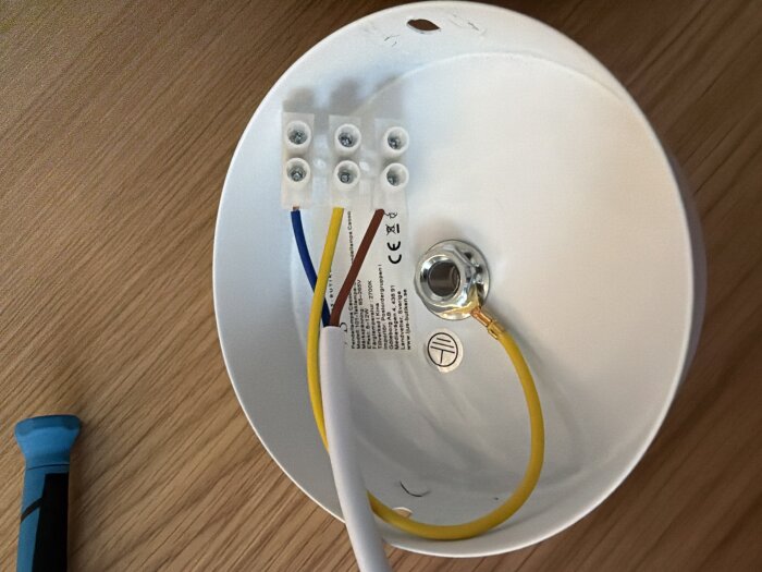 Vit lampplatta med anslutna kablar, inkluderande gul och brun kabel, samt skruvmejsel vid sidan.
