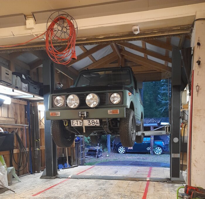 Grön Jeep lyfts upp på en billyft i ett garage med trädetaljer och verktyg synliga, och en mini Cooper står utanför i bakgrunden.