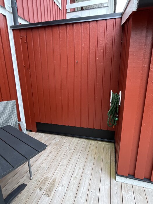 Röd trävägg vid altan med sittgrupp och grön trädgårdsslang på hållare.