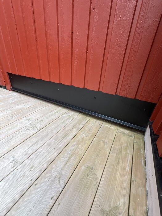 Nybyggd altan i trä intill en röd trävägg med svart plåtsockel.