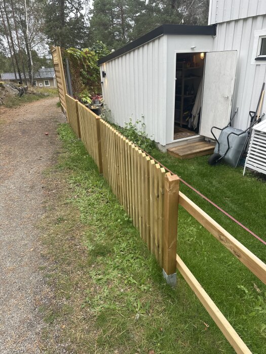 Trästaket med nya stolpar installerade i marken, beläget bredvid en vit byggnad. En gräsmatta och en grusgång syns bredvid staketet.