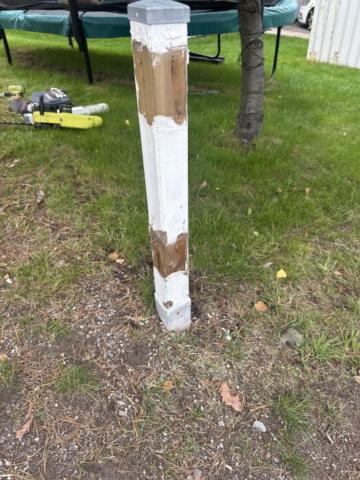 Sliten vitmålad staketstolpe fast i markspjut framför en trampolin; gräsmatta i bakgrunden.