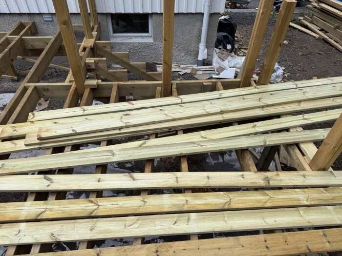 Trästruktur för altanbygge under uppförande, med staplade brädor nära en husvägg. Verktyg och byggmaterial syns i bakgrunden.