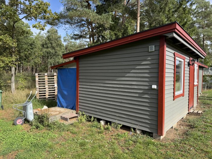 Grå vedbod med röda knutar, blå presenning täcker virkesförvaring. Ett skottkärra står bredvid, omgiven av grönska och träd i bakgrunden.