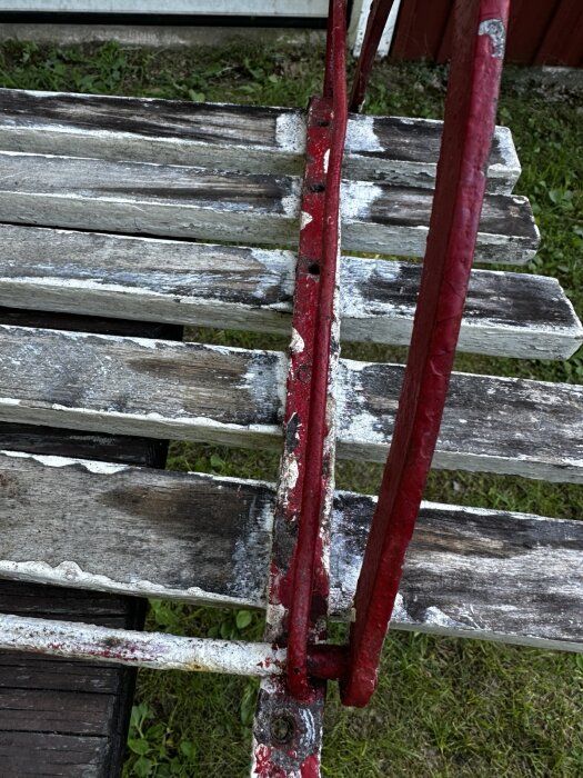 Sliten trädgårdssoffa med röda järndetaljer och flagnad vit färg på träplankor, stående på gräsmatta.