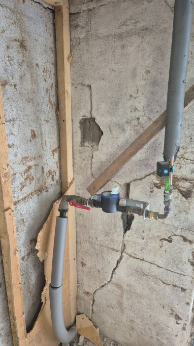 Vattenrör med avstängningsventiler och isolering på en grov betongvägg med sprickor och träreglar.