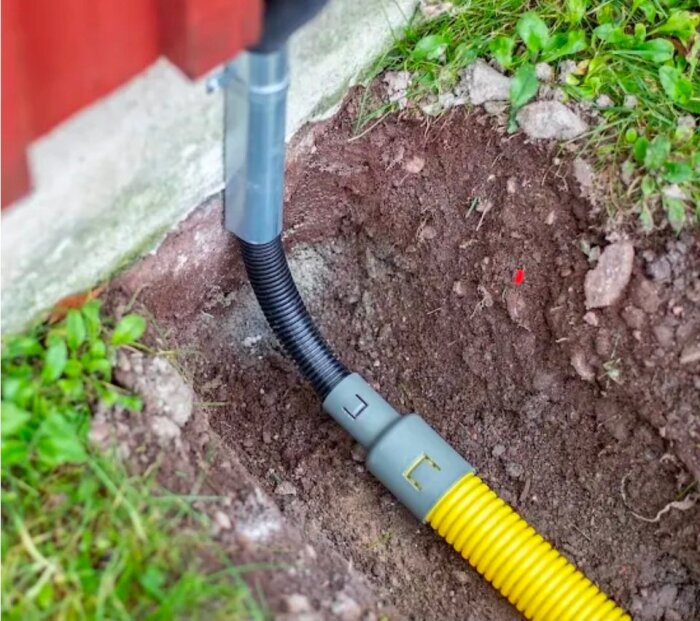Övergångsmuff med gul och svart kabel i marken intill en husvägg.
