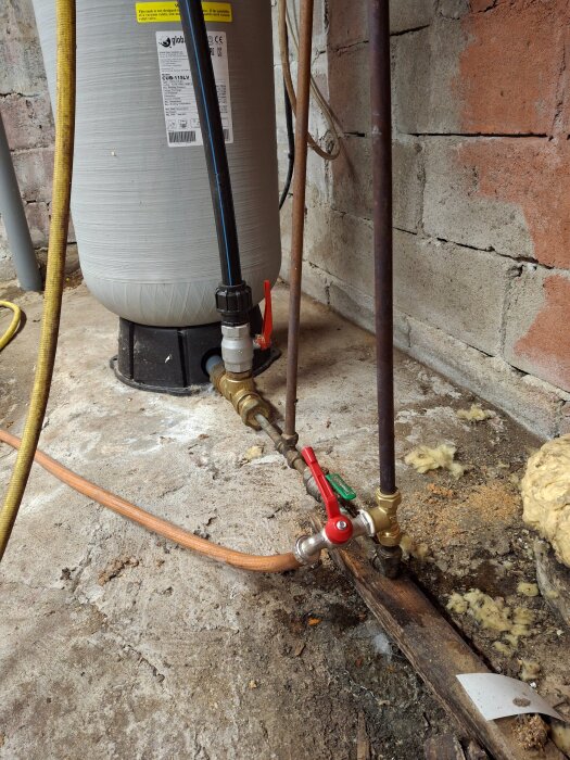 Trycktank med rör och ventiler i ett betongrum, visar ett komplext VVS-system mot en tegelvägg.