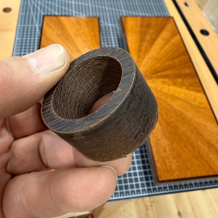 Närbild på en hand som håller i ett cylindriskt träobjekt, med två träluckor i bakgrunden på ett arbetsbord.
