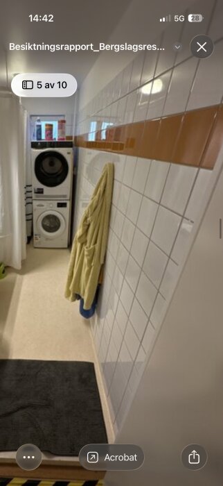 Tvättmaskin och torktumlare staplade intill kaklad vägg i ett badrum med handduk på väggen och plastmatta på golvet.