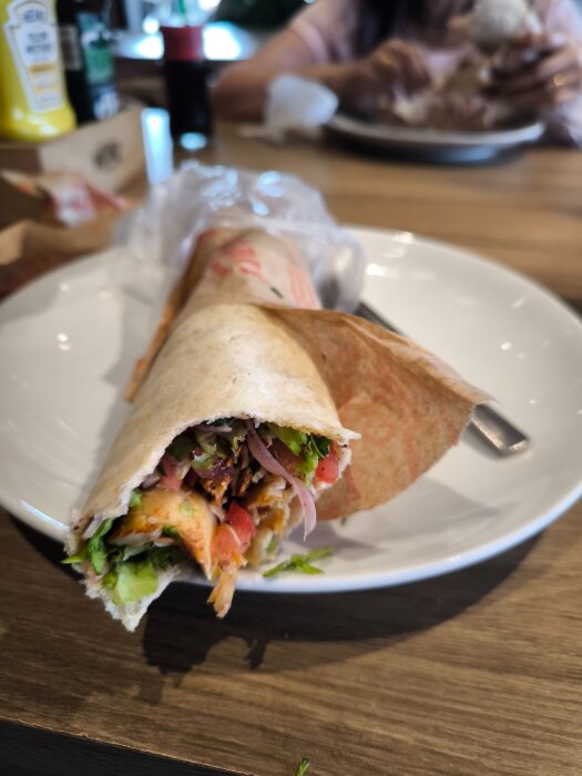 En halväten dönerkebab på en tallrik med sallad, tomater och lök. Bakgrunden visar en suddig person som äter.