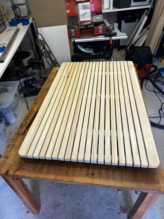 Ommålade träplankor redo för möbelrenovering ligger utlagda på ett träbord i en verkstad med verktyg och utrustning i bakgrunden.