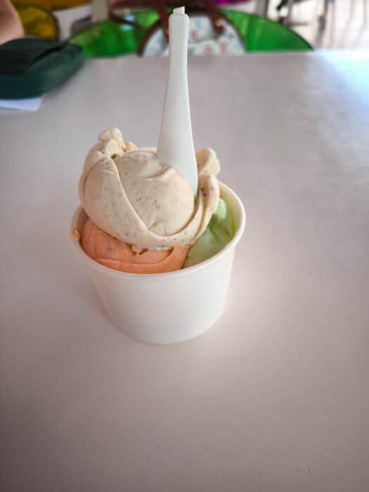 En vit kopp med tre kulor gelato i smakerna valnöt, persika och mint, serverad med en vit plastsked.