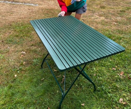 Grönt trädgårdsbord med metallunderrede, målat med linoljefärg, står på en gräsmatta. En person i färgglad tröja står vid bordet.
