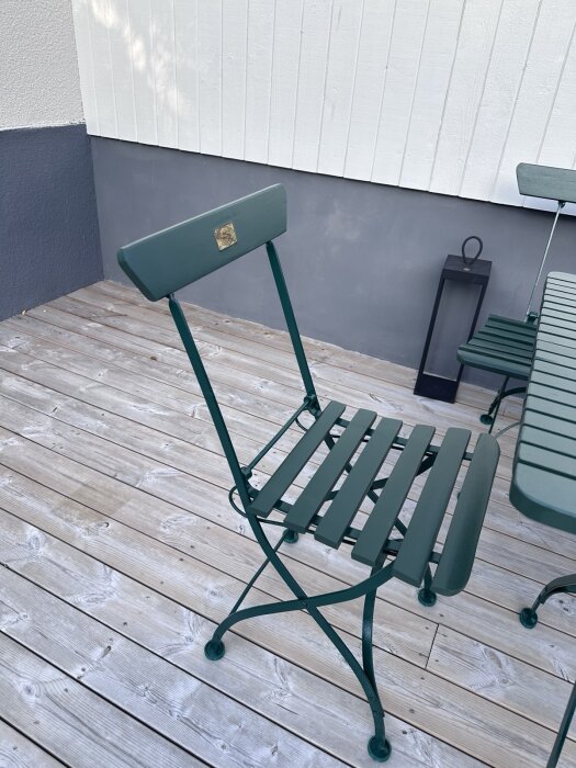 Mörkgrön metallstol med träinslag står på trädäck, lutad mot vitmålad vägg. En mörkgrön lykta syns i bakgrunden.