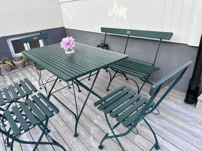 Grönmålad trädgårdsgrupp med bord, stolar och soffa på trädäck. En vas med rosa blomma står på bordet.