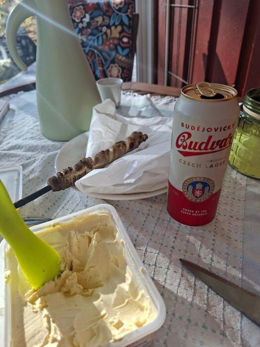 Öppnad ölburk, smörpaket med grön spatel, grillspett och vattenkanna på bord med duk i dagsljus.