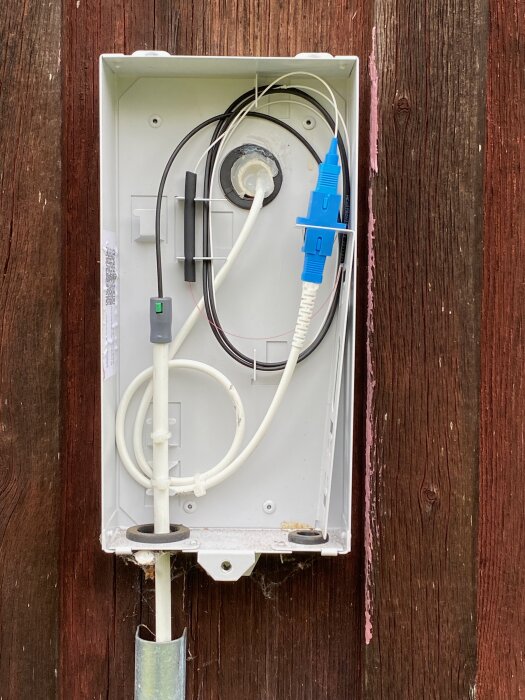Utomhusbox med fiberoptisk kabel och blå kontakt, monterad på trävägg. Vit fiberkabel leder till inomhusbox.