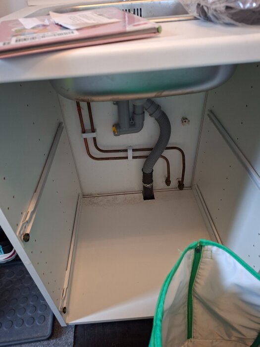 Under vasken i köksskåp, synliga rör och avlopp. Öppen förvaring, dokument och påse ovanpå diskbänken.