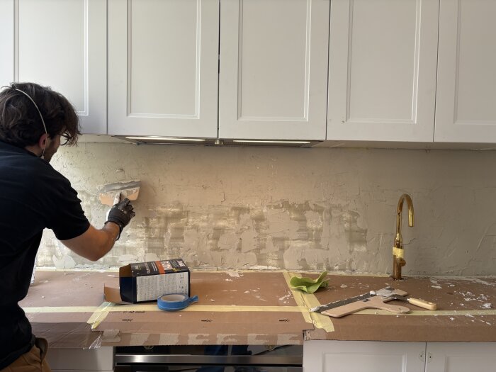 Person spacklar väggen i köket med cementbaserat spackel. Förpackning och verktyg syns på skyddad bänkskiva.