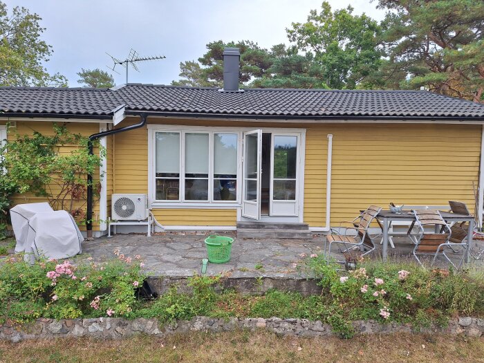 Gul sommarstuga med vita fönster och dörrar, stor stenlagd uteplats med trädgårdsmöbler, omgiven av träd och blommor.