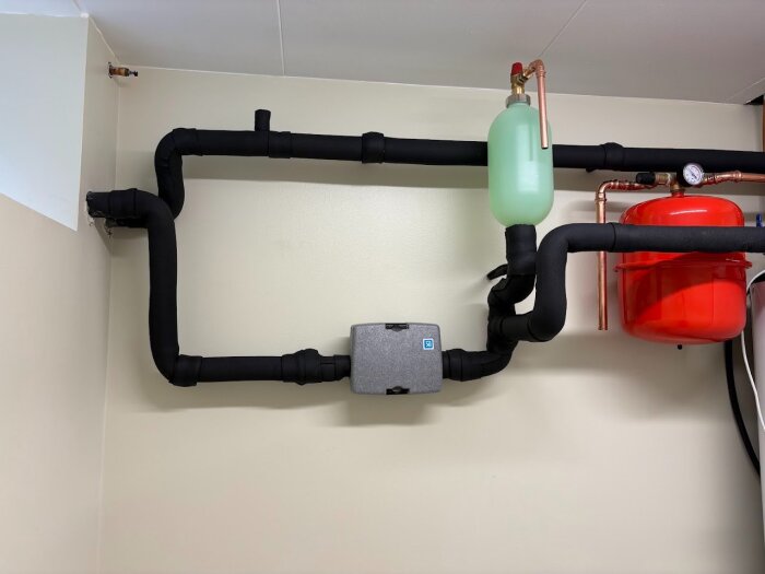 Bilden visar rörinstallation för en bergvärmepump med nivåbehållare monterad på utgående rör och isolerade rör. En grön behållare och röd tryckbehållare syns.