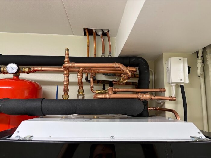Bergvärmepumpinstallation med kopparrör och tryckmätare, svarta isolerade rör, och en röd cistern, där nivåkärlet är på utgående bärrör.