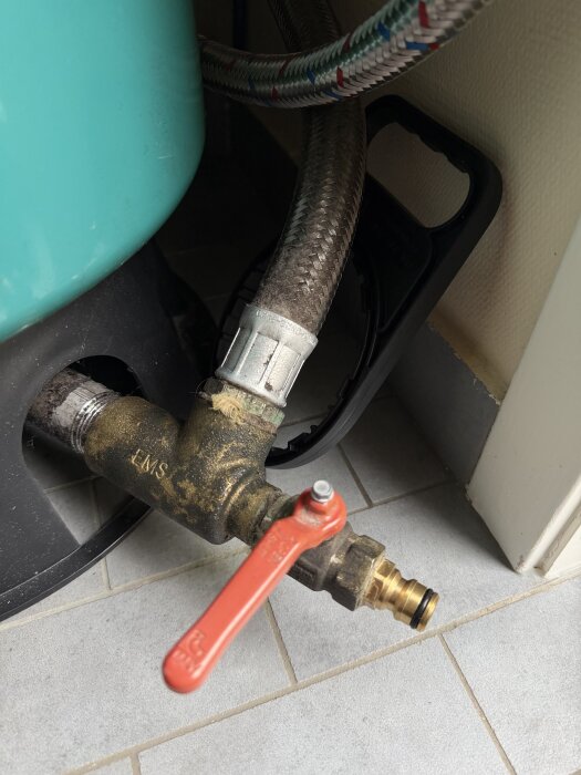 Hydropressventil med röd vred och snabbfäste för trädgårdsslang. Strukturerad anslutning och metallkopplingar synliga på klinkergolv.