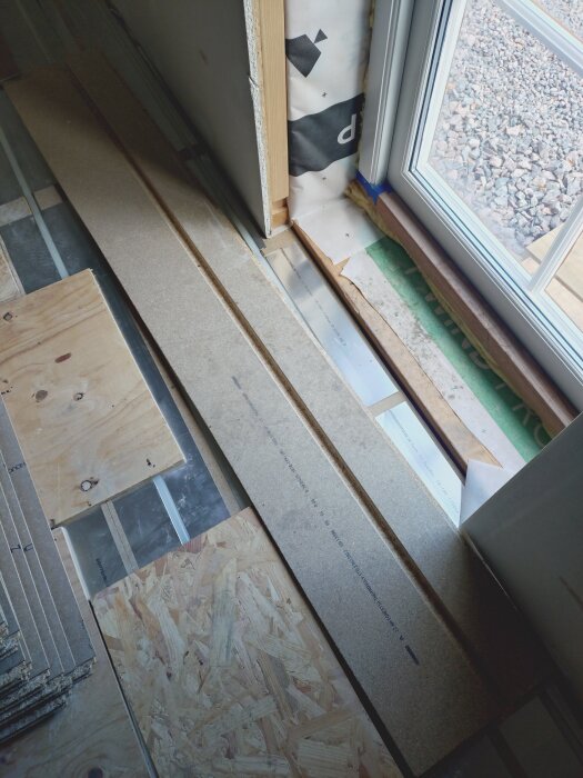 Golv med träskivor och springor synliga vid ett fönster, där golvvärmeslingor lagts och isolering saknas i bjälklagets fack.