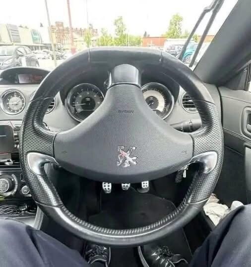 Förarplats med ratt i en bil, instrumentpanel med hastighetsmätare och radio synliga, bilar och byggnader utanför fönstret.
