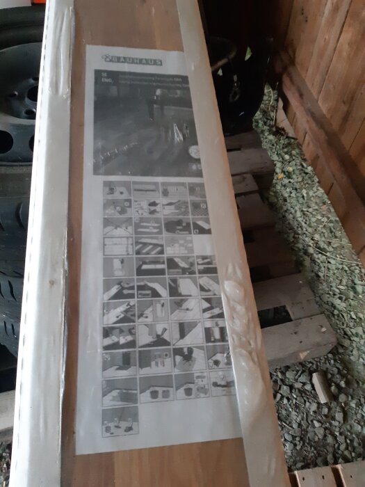 Paket med Bauhaus ekparkett standard liggande på trähylla i carport, delvis synlig bruksanvisning på ovansidan.