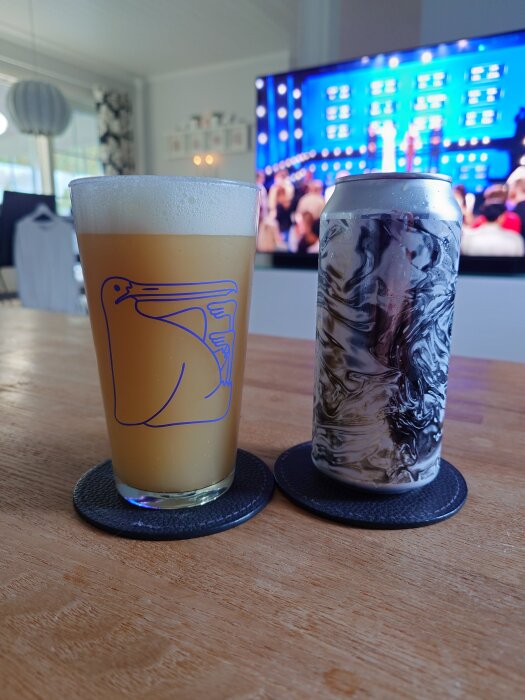 Ett glas öl med logotyp och en abstrakt mönstrad dryckesburk på ett bord framför en TV.