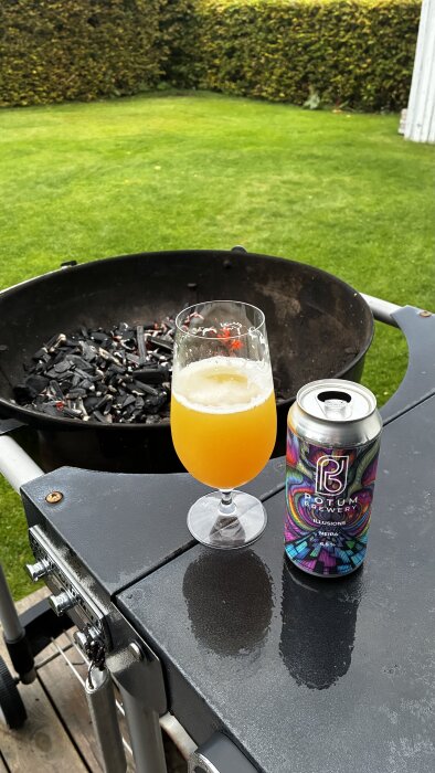 Glas med orange öl och burk Potum Illusions NEIPA på ett bord vid en grill med glödande kol, gräsmatta i bakgrunden.