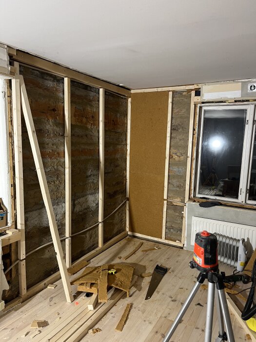 Byggprojekt i rum med reglar och obs-skivor monterade på väggar; verktyg och material på golvet, och en laser på stativ vid sidan.