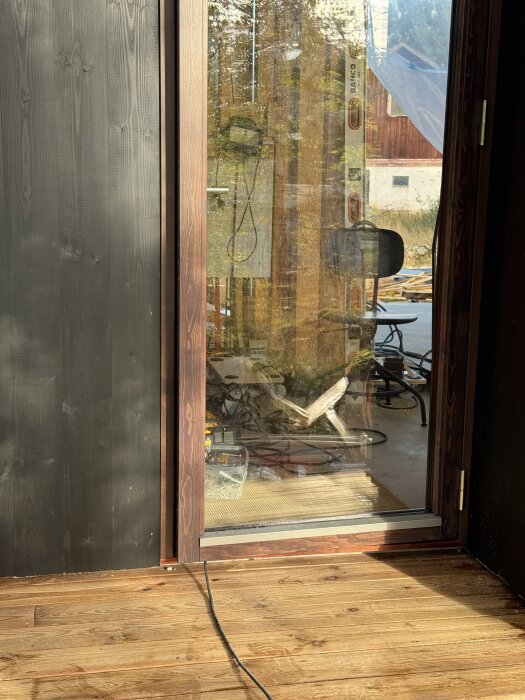 Trädörr med glasdelar mot terrass, synliga byggverktyg reflekteras i glasset, plankgolv och svart fasadpanel.