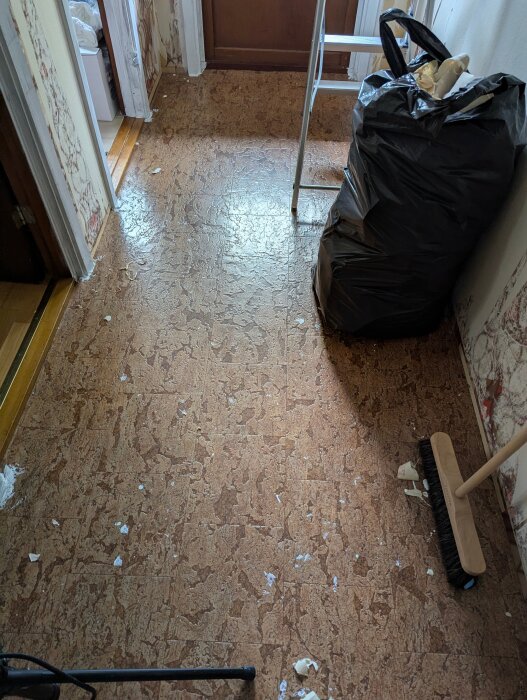 Hall med brunaktig retro matta, skräp på golvet, sopborste och svarta sopsäckar synliga. En stege står mot väggen vid dörren.