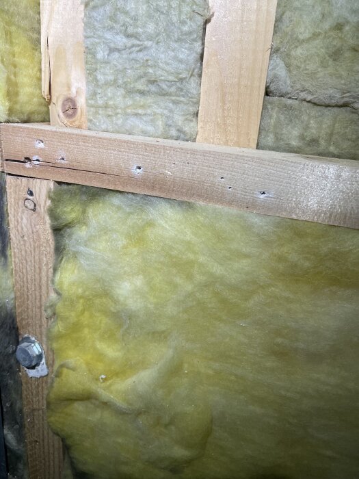 Gul isolering av glasfiber synlig bakom träreglar i en väggkonstruktion med synliga skruvhål och en metalldetalj till vänster.
