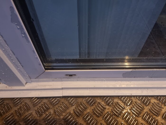 Vattenstänk på ett glasfönster intill en dörrkarm och ett golv av räfflad metall.