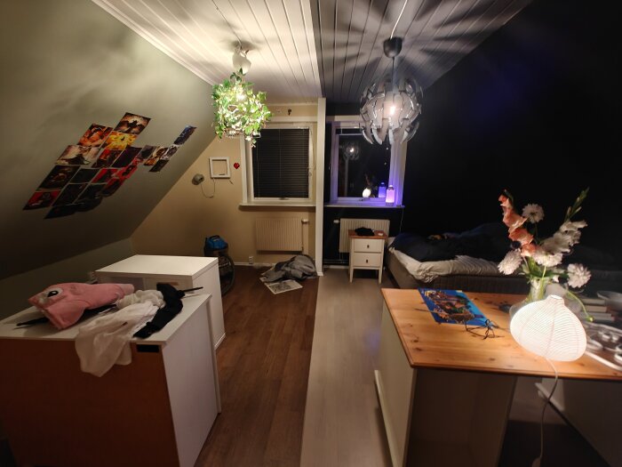Sovrum med lofttak, säng och skrivbord. Vägg med fotografier, växtlampa och nattduksbord. Bord med lampa och blommor.