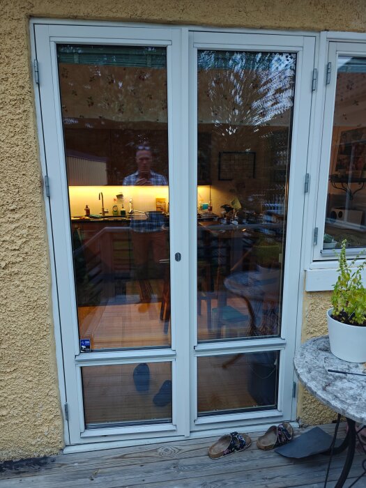 Glasdörrar med reflektion av man och kök, vägg i gult puts. Bord med krukväxt och skor på trädäck.