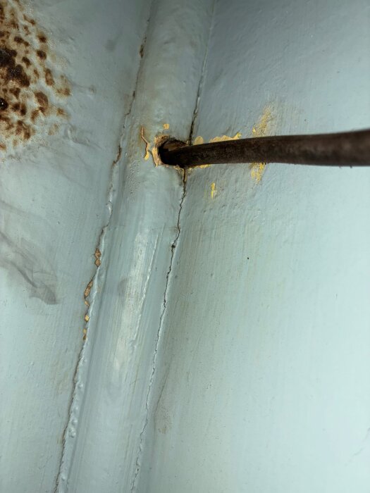 Rostig metallstång genom ett hål i en blåmålad yta med sprickad färg och rostfläckar intill.