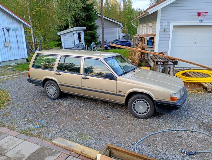 Beige Volvo kombi parkerad på en stenlagd yta, omgiven av träd och små byggnader, med verktyg och material i närheten.