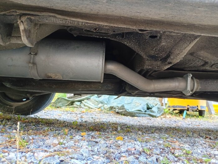 Nytt avgassystem på en bil sett underifrån, monterat från katalysatorn bakåt, med detaljer som märke och utformning. Grusig mark syns under bilen.
