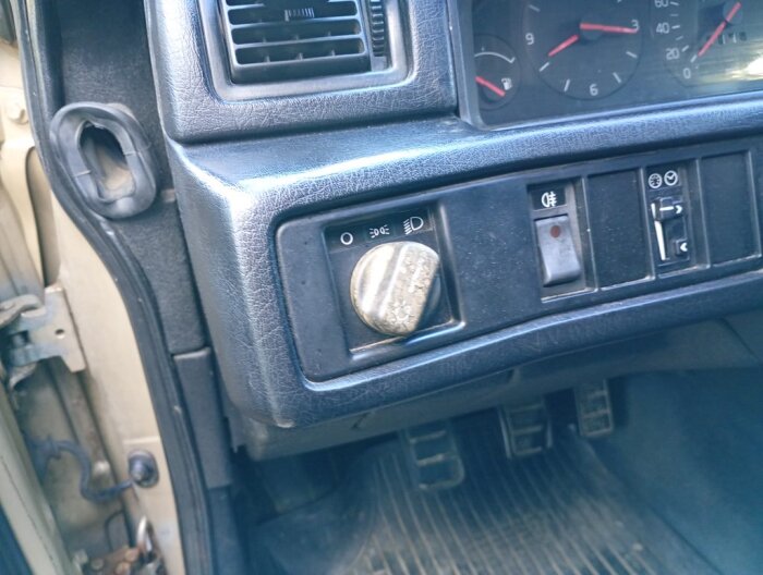 Slitet vred för belysning i en gammal Volvo, blå instrumentpanel och fotutrymme i bilen synliga, samt dörrens gummilist och instrumentbräda.