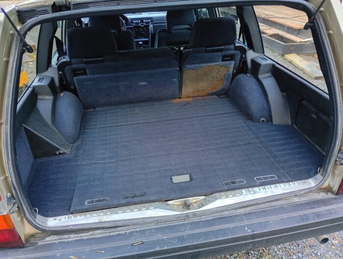 Bild av bilens bagageutrymme med nedfällt baksäte, synliga slitage och original gummimatta från Volvo, inuti en äldre bilmodell.