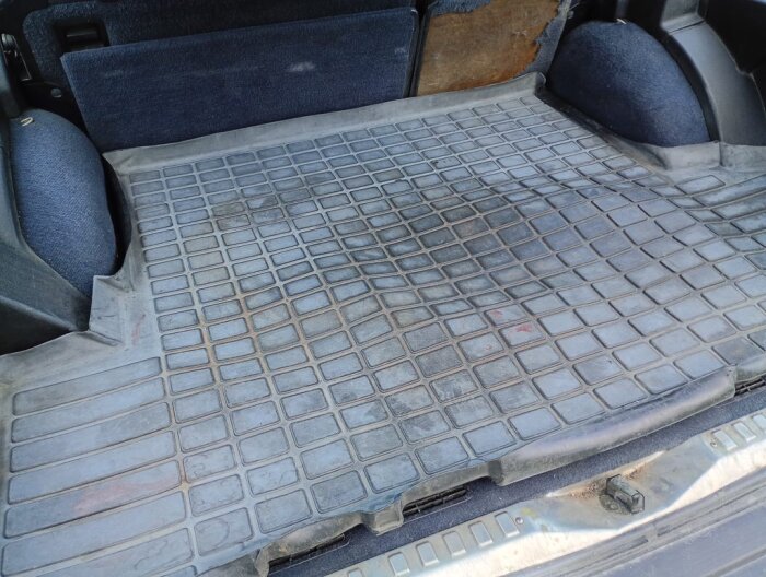 Bilden visar en delvis smutsig original Volvo-gummimatta i baksätets lastutrymme, omgiven av blå klädsel och plastpaneler.