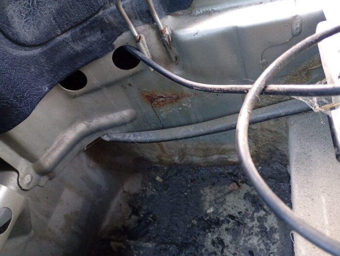 Bilden visar en bils bagageutrymme med spår av rost på metallen nära två kablar, smutsig ytbeläggning och en plastpanel ovanför.